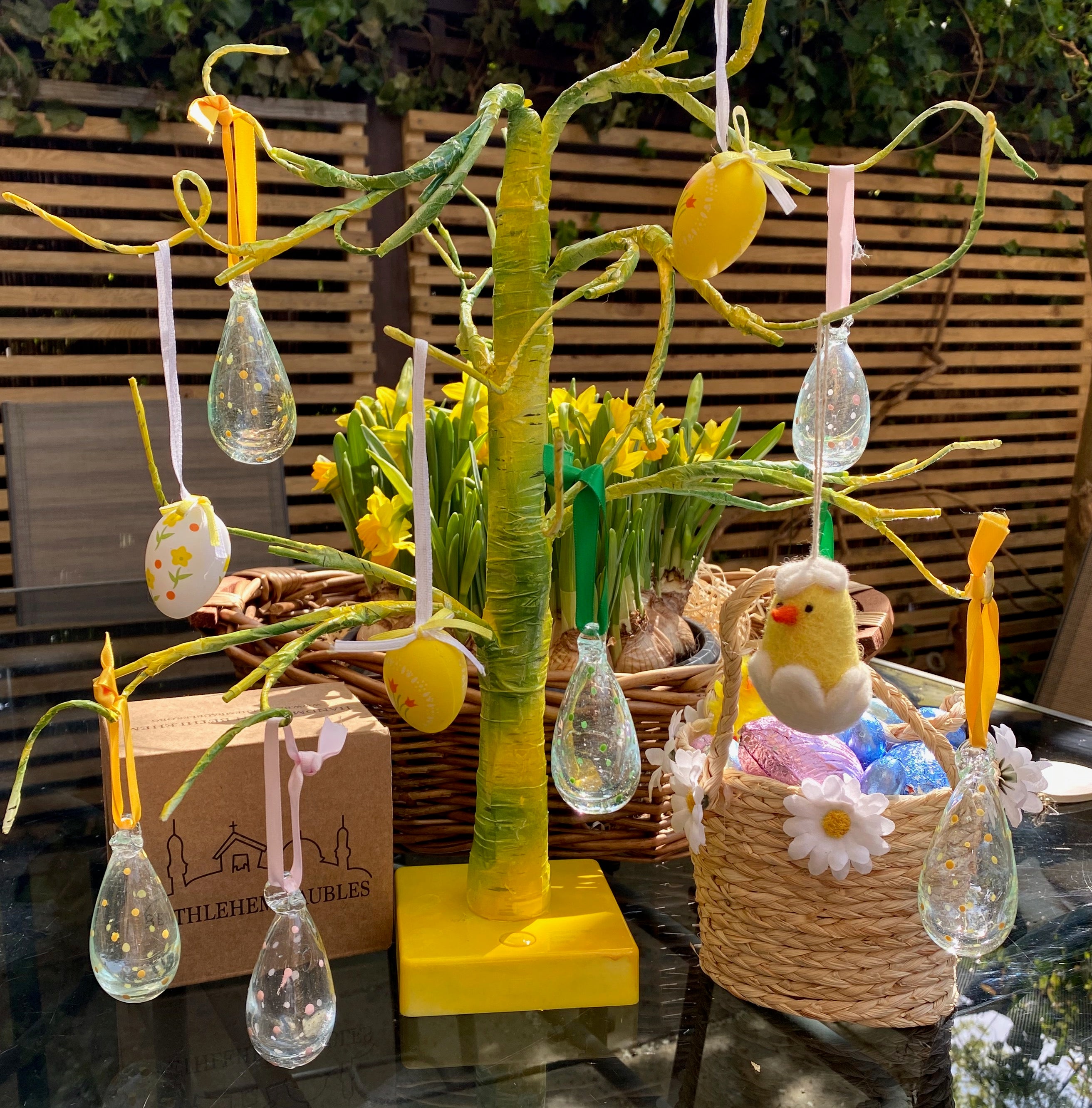 Decorative Easter tree with hanging lights, a chick, and flowers on a table outdoors.
